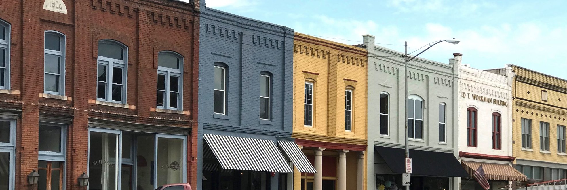 A row of colorful historic brick buildings with large windows and striped awnings lines a small-town street under a blue sky. A pink truck is parked in front.