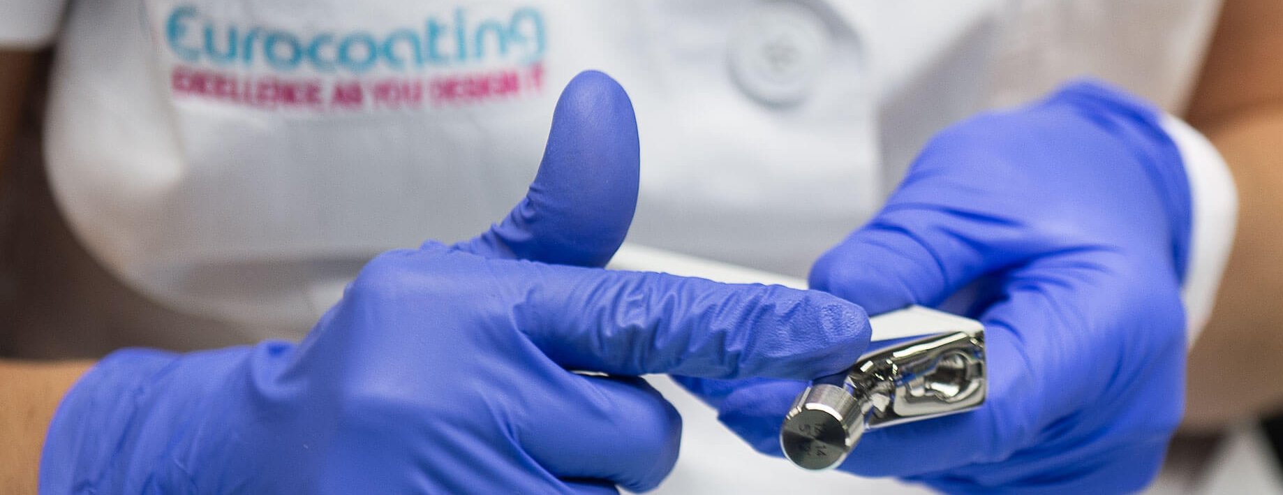 A person wearing purple gloves holds a shiny metal dental tool. Part of their white coat with the logo "Eurocoating" is visible in the background.