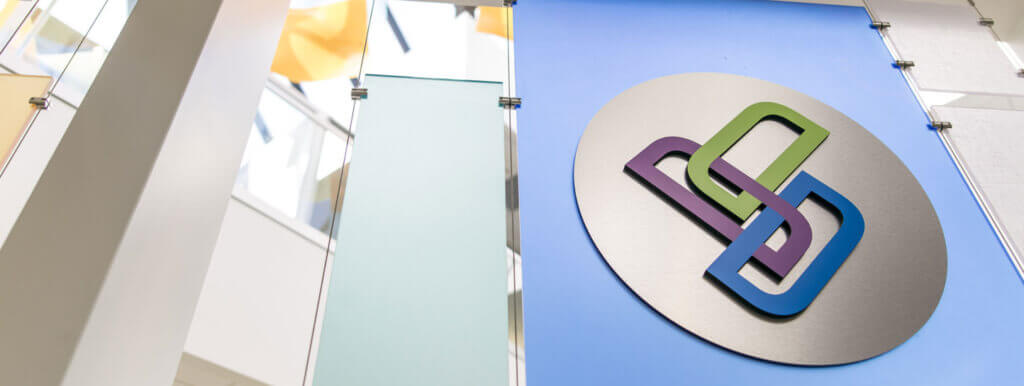A modern office interior featuring a large logo with interlocking green, purple, and blue shapes mounted on a blue and gray wall panel, with glass and sunlight in the background.