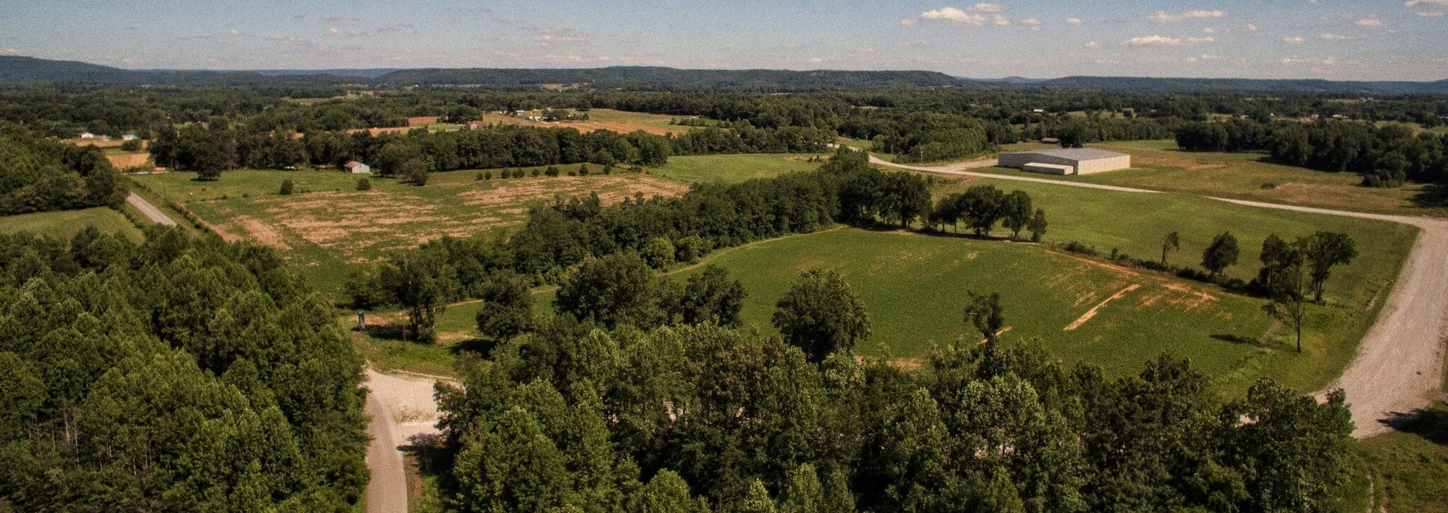 Aerial view of a rural landscape with green fields, clusters of trees, a long road, scattered buildings, and a large barn-like structure under a blue sky with scattered clouds.