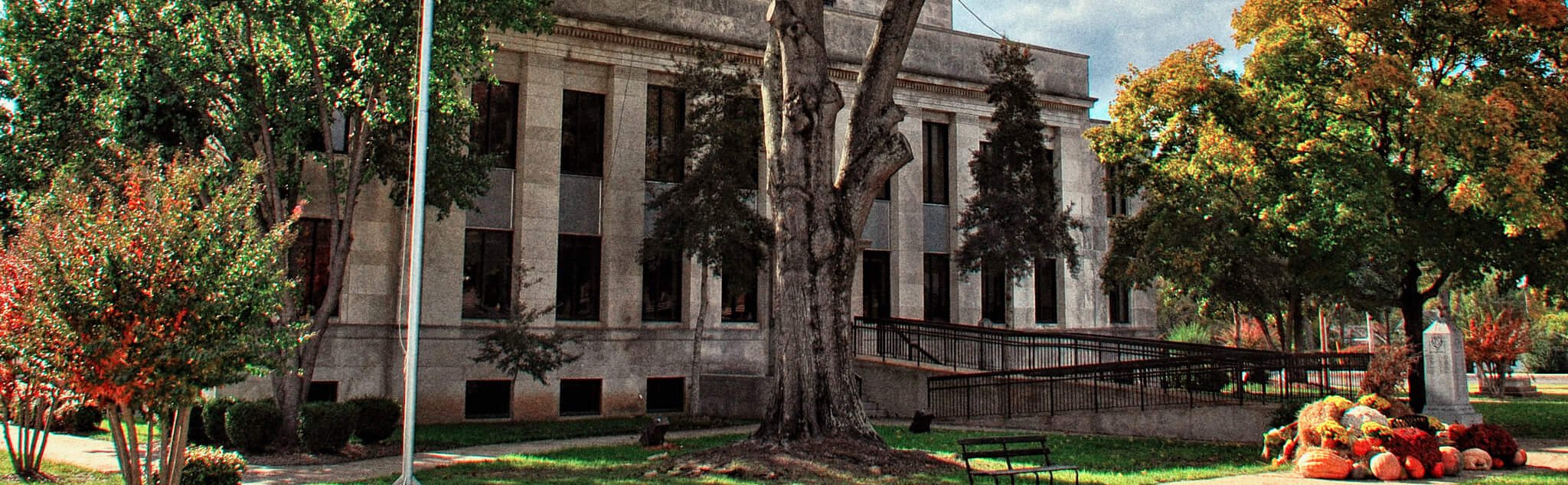 A historic stone building with tall windows is surrounded by trees and a lawn. A flagpole stands in front, and a pile of pumpkins and hay bales is arranged near a bench and a sidewalk.