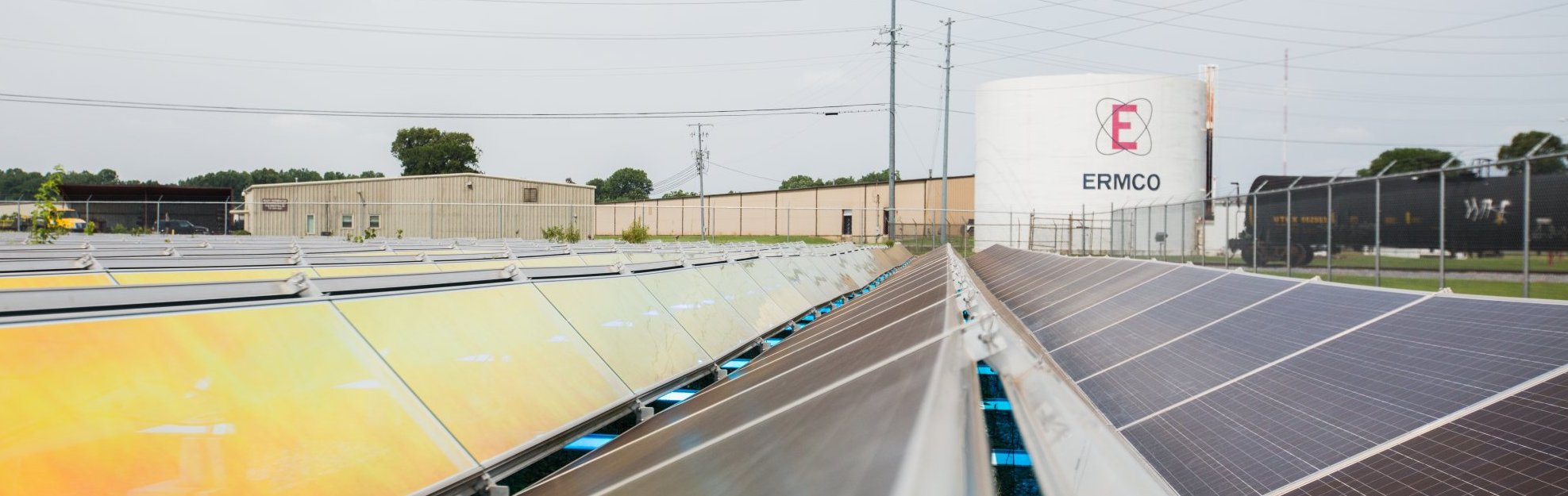 Rows of solar panels are installed outdoors near an industrial building and a large white storage tank labeled “ERMCO” under a cloudy sky, with power lines and trees in the background.