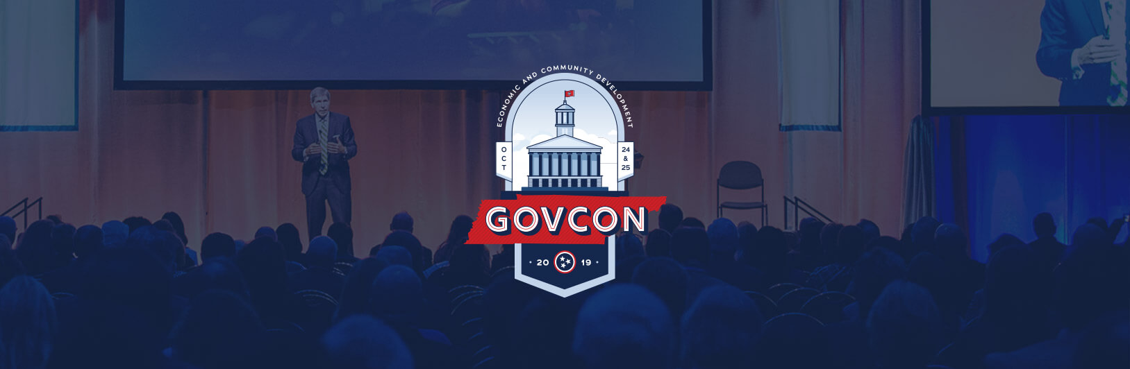 A speaker stands on stage in front of an audience at a conference. The GOVCON logo with a government building graphic is centered, with large screens and stage lighting visible in the background.