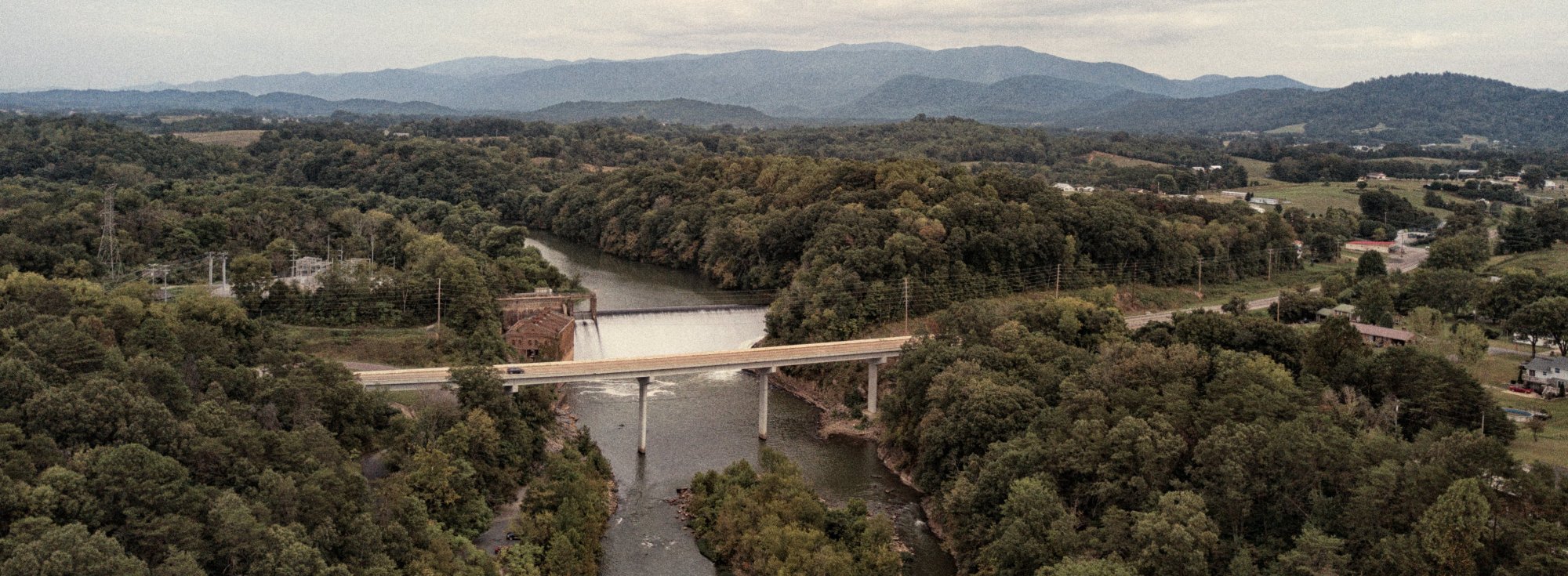 Aerial view of a bridge crossing over a river surrounded by dense green trees, with a dam nearby and mountains visible in the hazy background under a cloudy sky.
