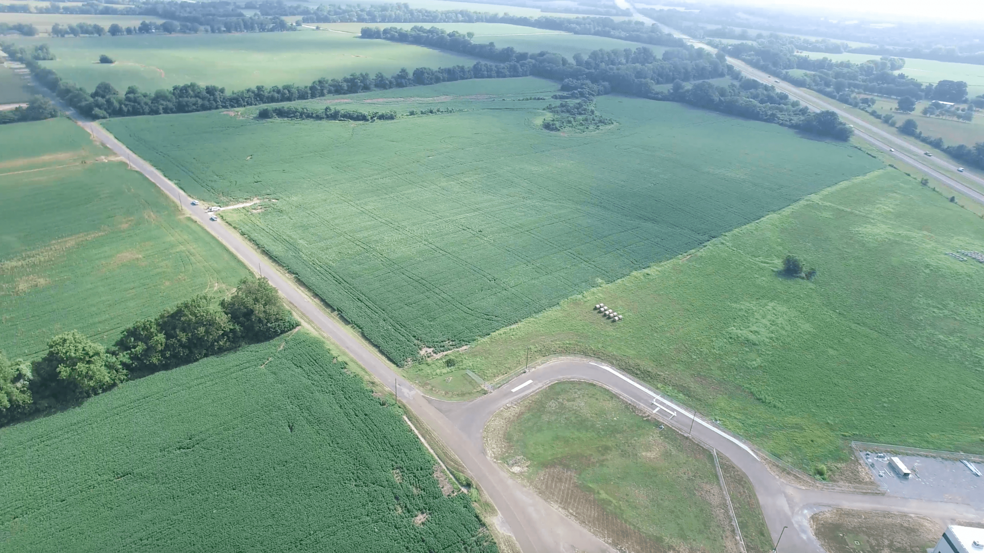 Aerial view of green farmland divided by roads, with fields, trees, and a small clearing. A paved circular area is visible in the foreground, and the landscape stretches out to the horizon under a light sky.