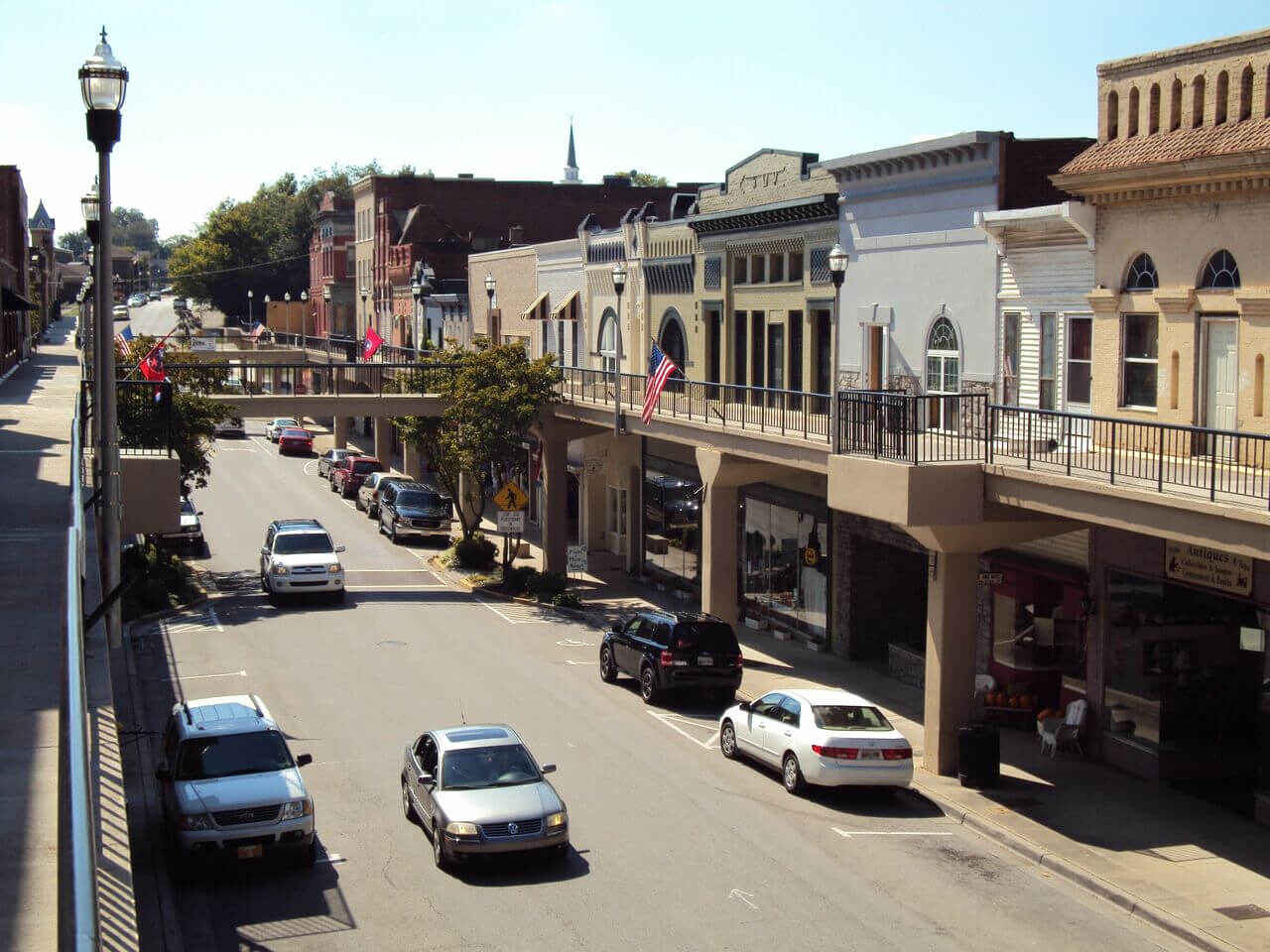 A small town street with parked cars, storefronts, and a pedestrian walkway above the road. American flags hang from the buildings, and a church steeple is visible in the background under a clear sky.
