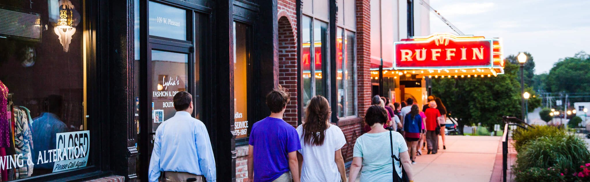 People walk along a sidewalk toward the brightly lit marquee of the Ruffin theater in a small town. Storefronts line the street, and the evening sky is visible in the background.