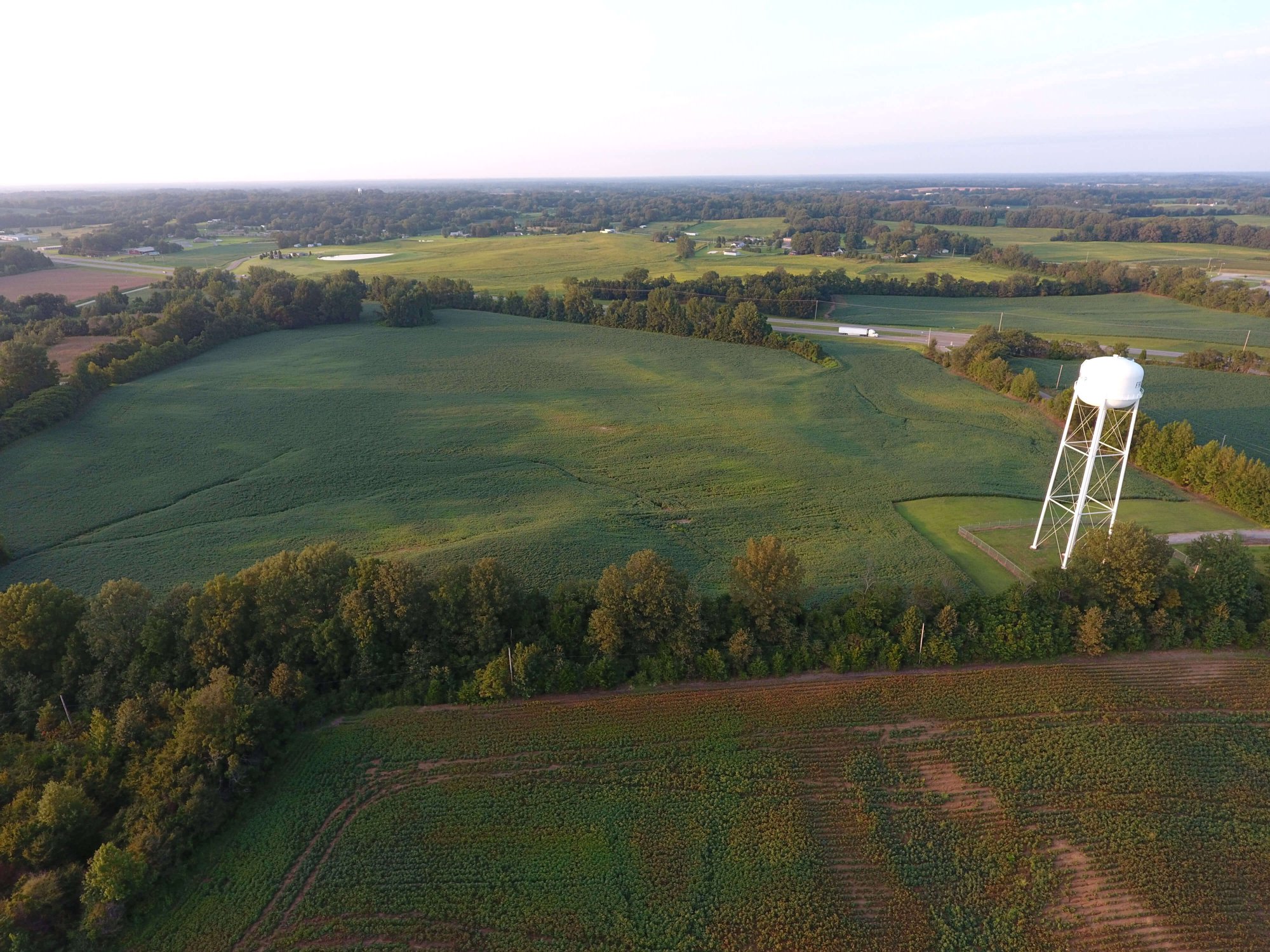 Aerial view of green farmland with fields, patches of trees, and a tall white water tower near a road, under a partly cloudy sky during daylight.