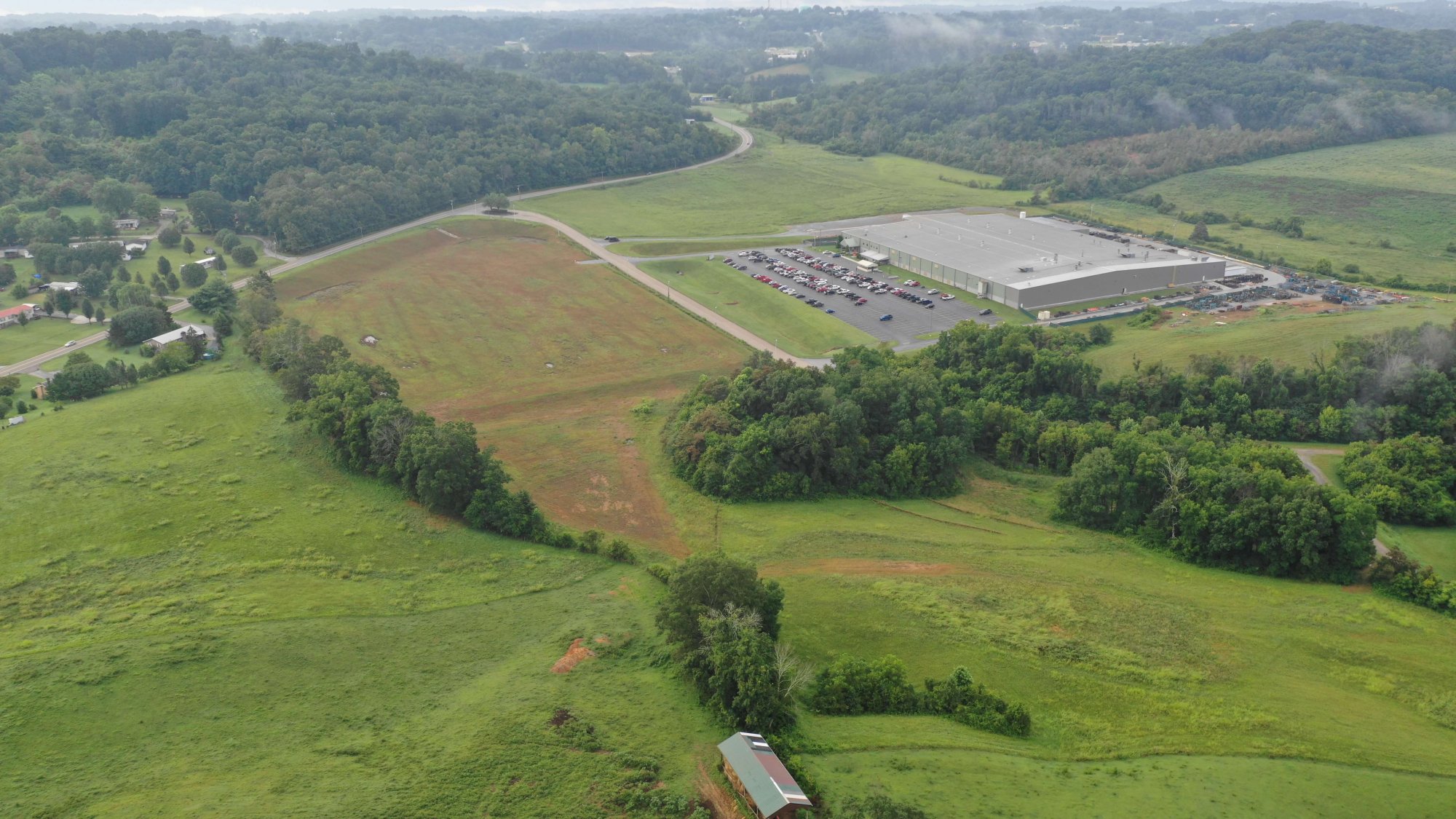 Aerial view of a large warehouse building with a parking lot, surrounded by open grassy fields, trees, and scattered houses; a winding road passes through the green landscape under a cloudy sky.