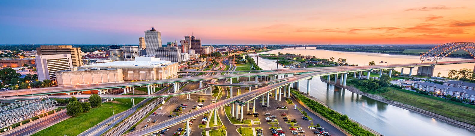 Aerial view of downtown Memphis, Tennessee at sunset, showing city buildings, highways crossing over the Mississippi River, and a bridge with the sky glowing orange and pink.