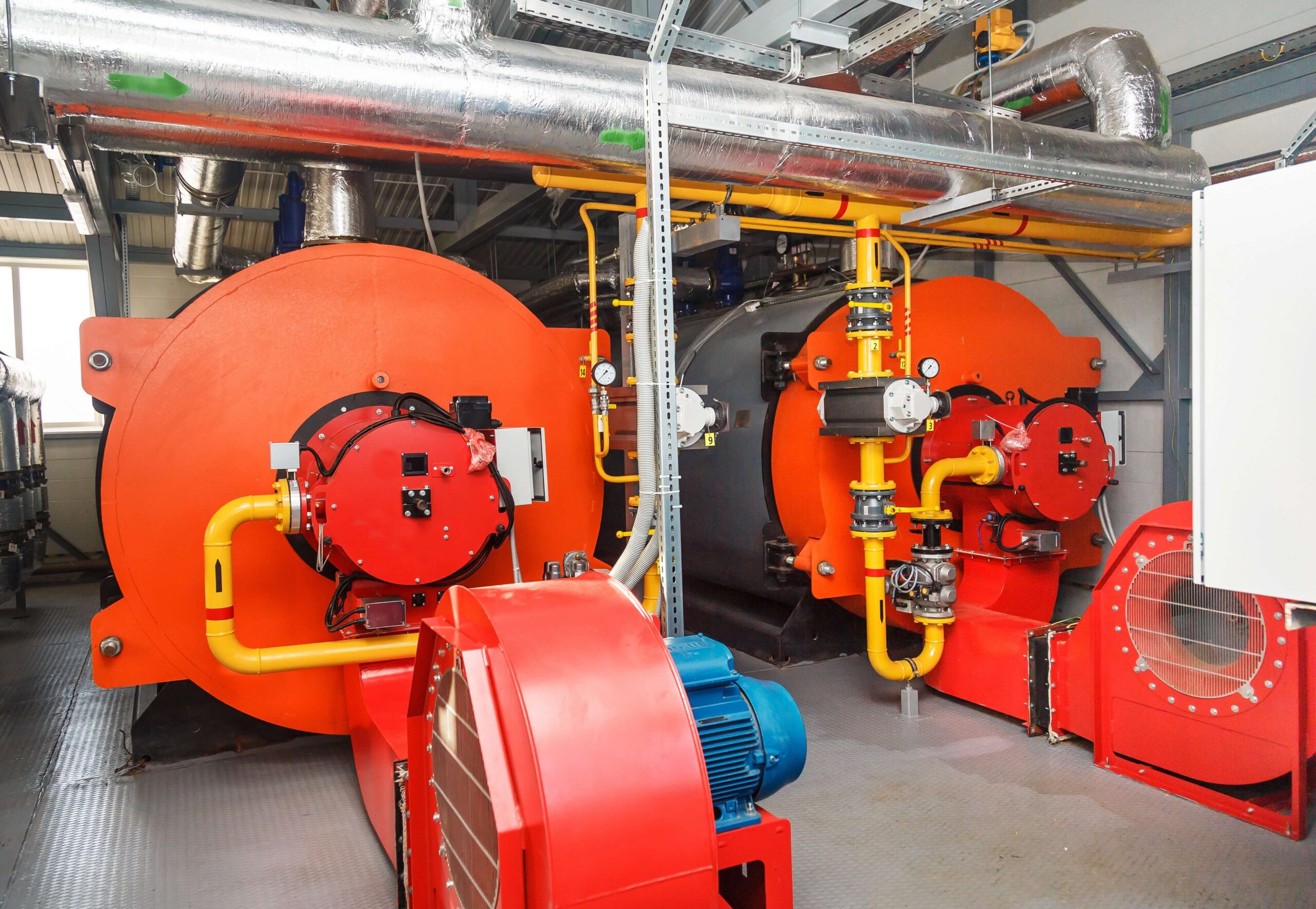 Two large, orange industrial boilers with yellow pipes and control panels in a mechanical room, surrounded by metal ducts and various machinery.
