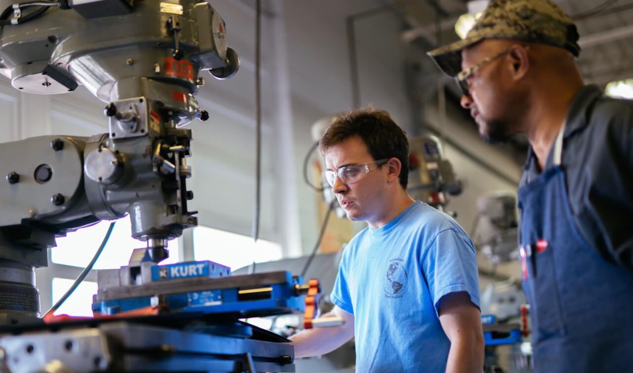 Two men wearing safety glasses work with a large metal milling machine in a workshop. One man, in a blue shirt, closely observes the machine, while the other, in a cap and apron, stands beside him.