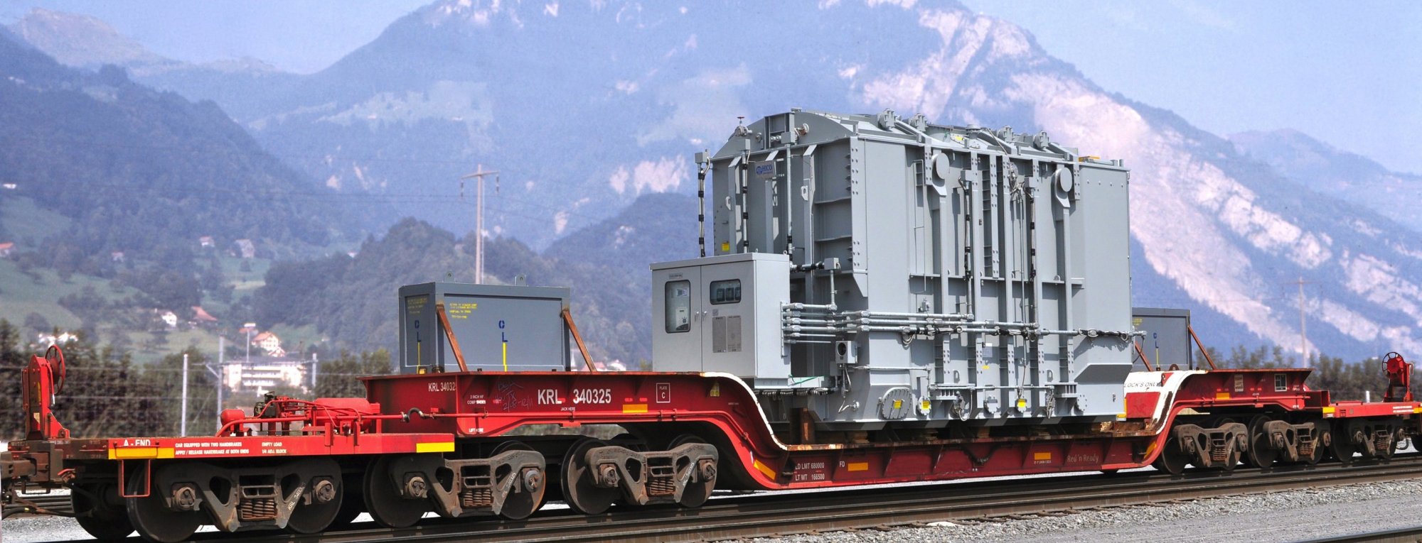 A large industrial transformer is securely mounted on a red flatbed railcar, traveling on train tracks with mountains and a partly cloudy sky in the background.