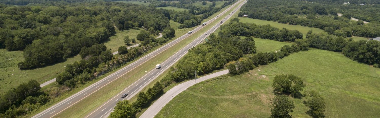 Aerial view of a divided highway running through a green, rural landscape with trees and open fields on both sides and several vehicles traveling in both directions.
