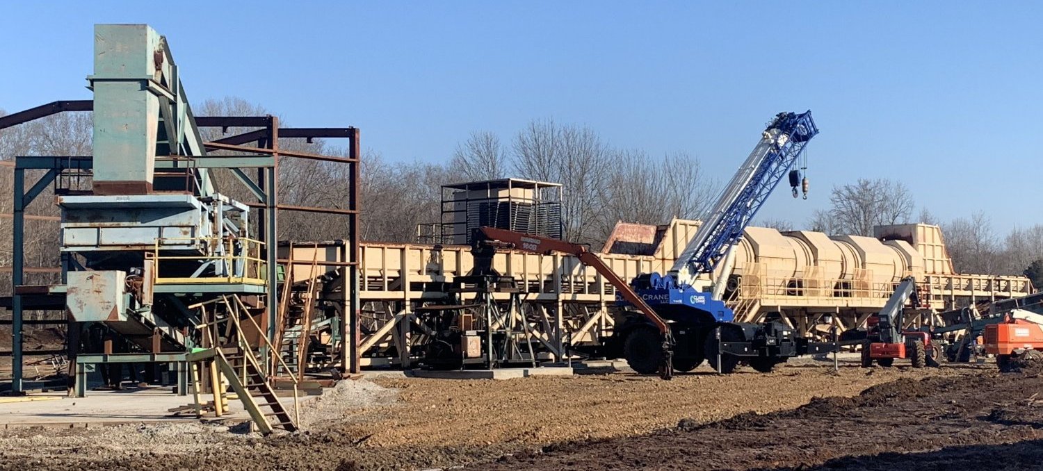 A construction site with large industrial machinery, including a blue crane and several tan-colored conveyor structures, sits on dirt ground with leafless trees and a clear blue sky in the background.