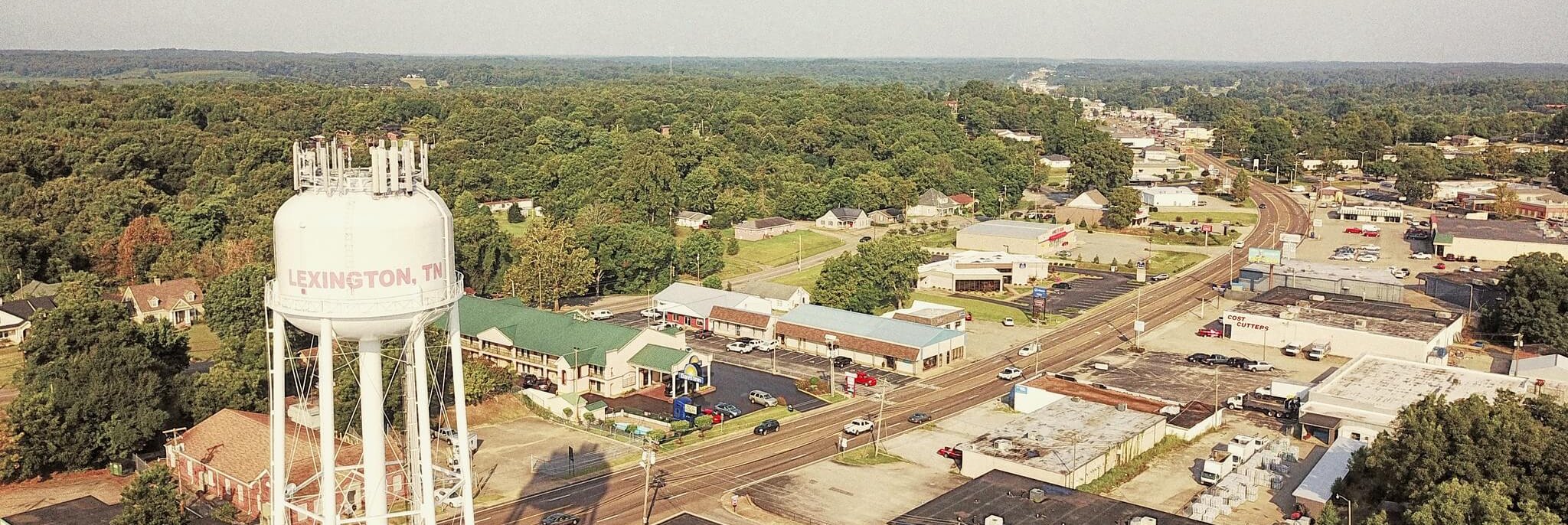 Aerial view of a small town with a white water tower labeled "LEXINGTON, TN," surrounded by trees, houses, and commercial buildings along a curving road under a clear sky.