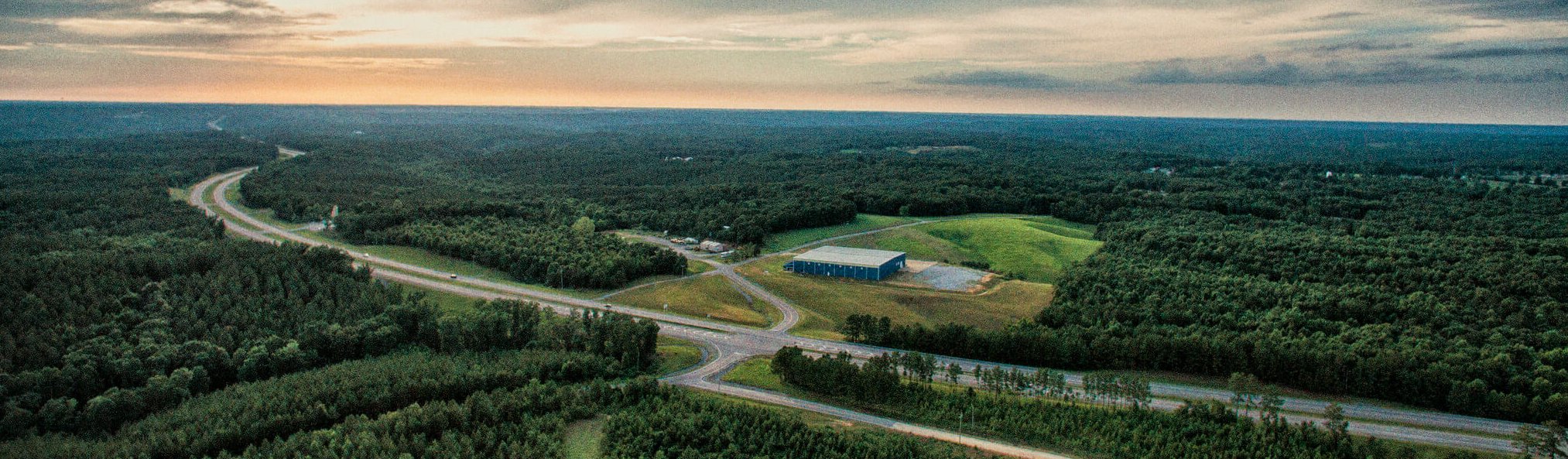 Aerial view of a highway intersection surrounded by dense green forest, with a large building and open grassy area near the road, under a cloudy sunset sky.