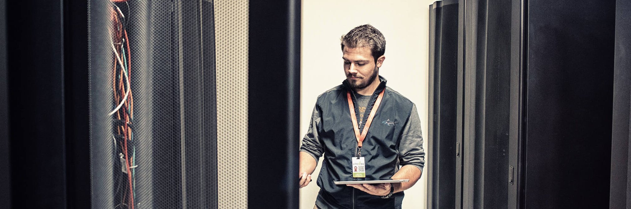 A man holding a laptop stands between server racks in a data center, inspecting equipment and wearing an employee badge on a lanyard.