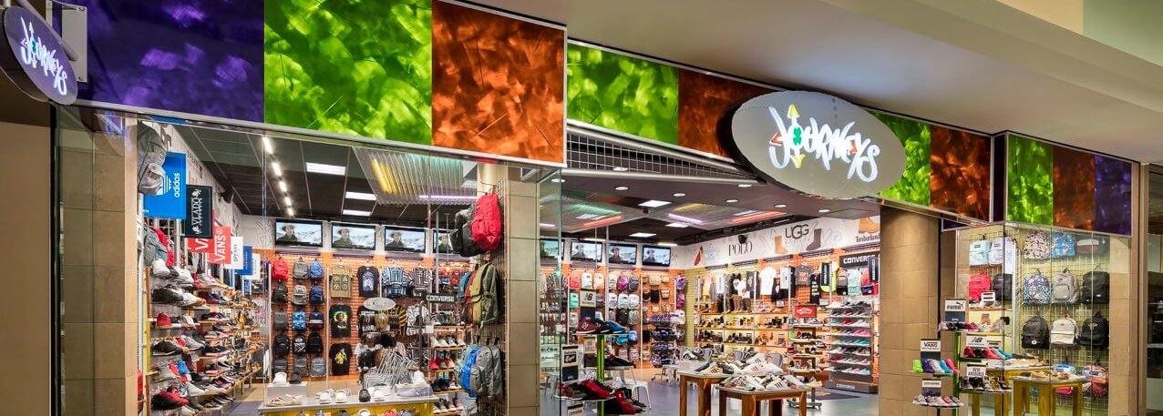 A colorful shoe store with glass walls displays a variety of sneakers and footwear on shelves. Bright signage and multicolored panels decorate the storefront, and multiple TVs line the top inside the store.