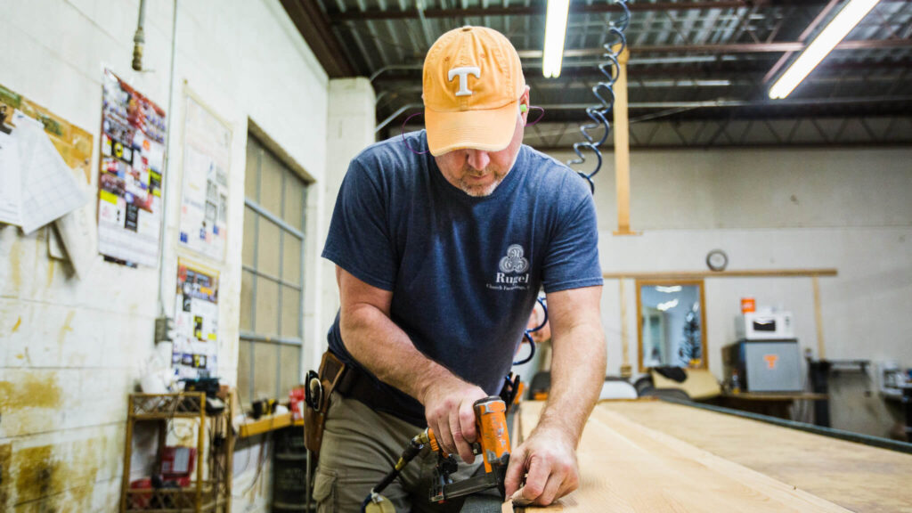 A man wearing an orange cap and blue shirt uses a power tool to work on a wooden board in a workshop with tools, posters, and industrial lighting in the background.