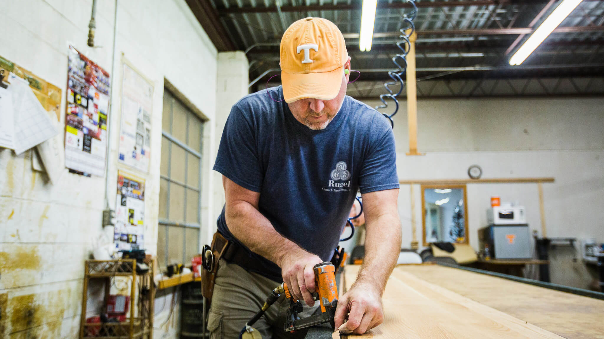 A man wearing an orange cap and blue shirt uses a power tool to work on a wooden board in a workshop with tools, posters, and industrial lighting in the background.