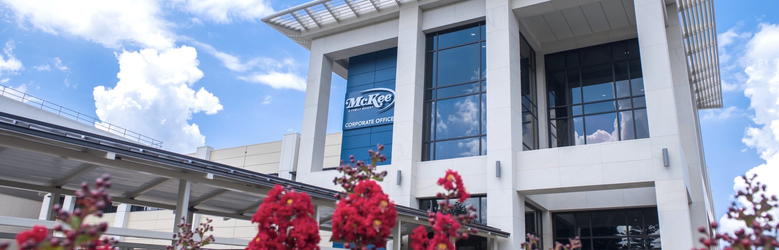Modern white office building with large glass windows, a blue sign reading "McKee Corporate Office," and red flowering plants in the foreground under a bright, partly cloudy sky.