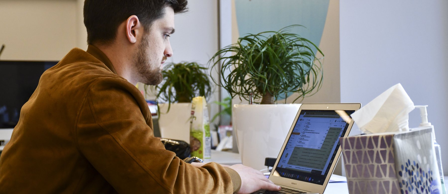 A man in a brown jacket works on a laptop at a desk, with potted plants, a tissue box, and office supplies in the background.