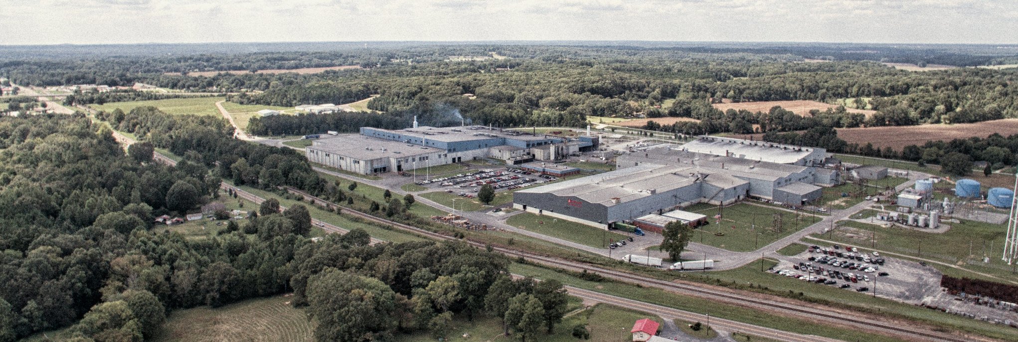 Aerial view of a large industrial facility surrounded by parking lots, trees, and fields, with a railway track in the foreground and a forested landscape in the background under a cloudy sky.