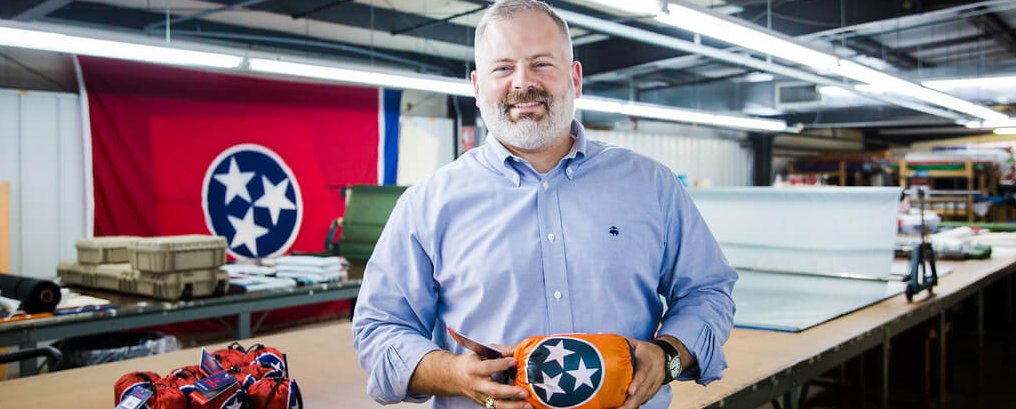 A smiling man with a beard stands in a workshop holding an item with the Tennessee state flag symbol; a large Tennessee flag hangs on the wall behind him, and worktables with supplies are visible in the background.