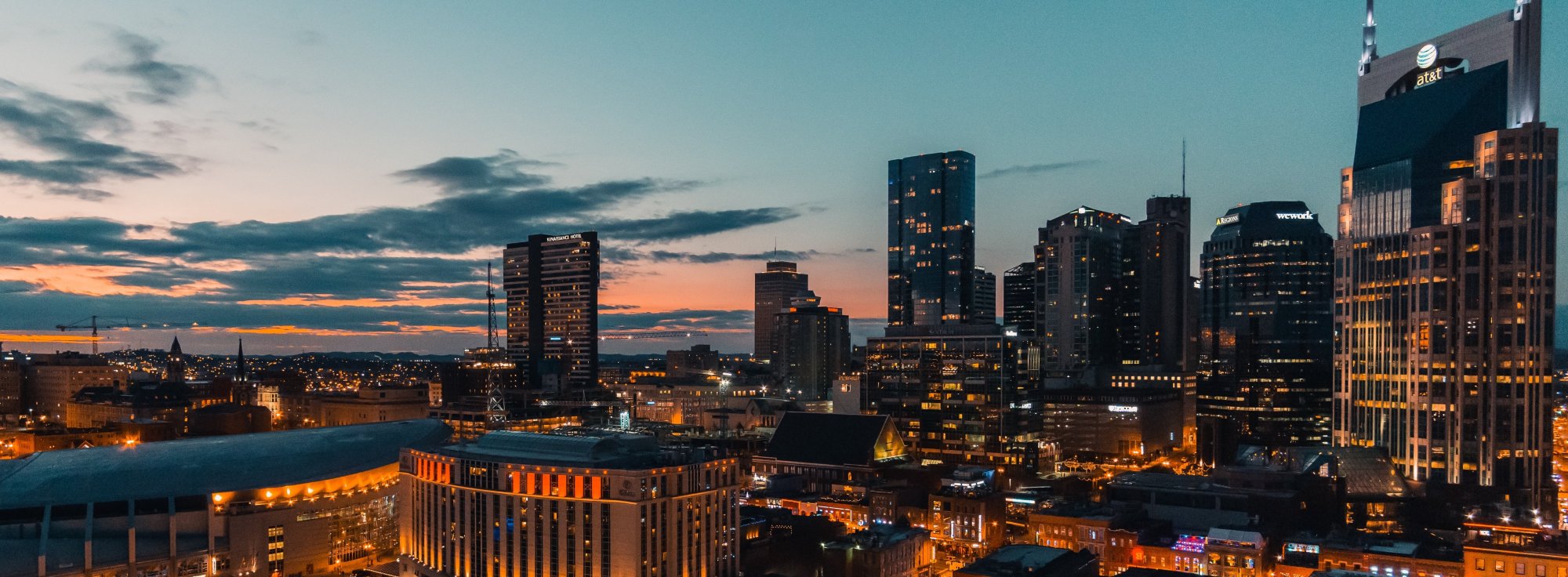 A city skyline at dusk with tall, modern buildings lit up against a colorful sunset sky, featuring a blend of blue, orange, and yellow hues.