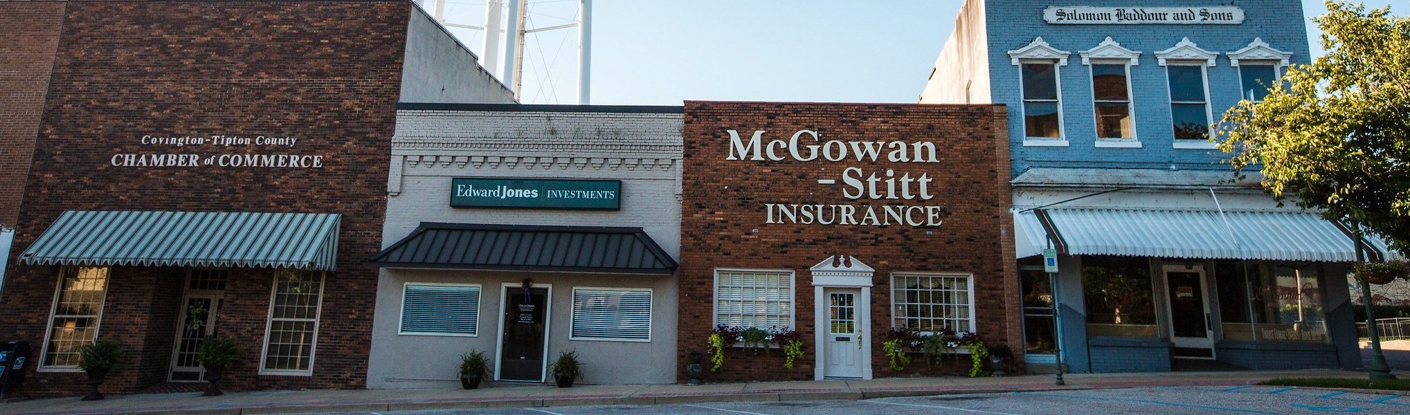 A row of three small-town brick buildings houses a chamber of commerce, an investment office, and an insurance agency, each with distinct signage and striped awnings, on a quiet street with parked cars.
