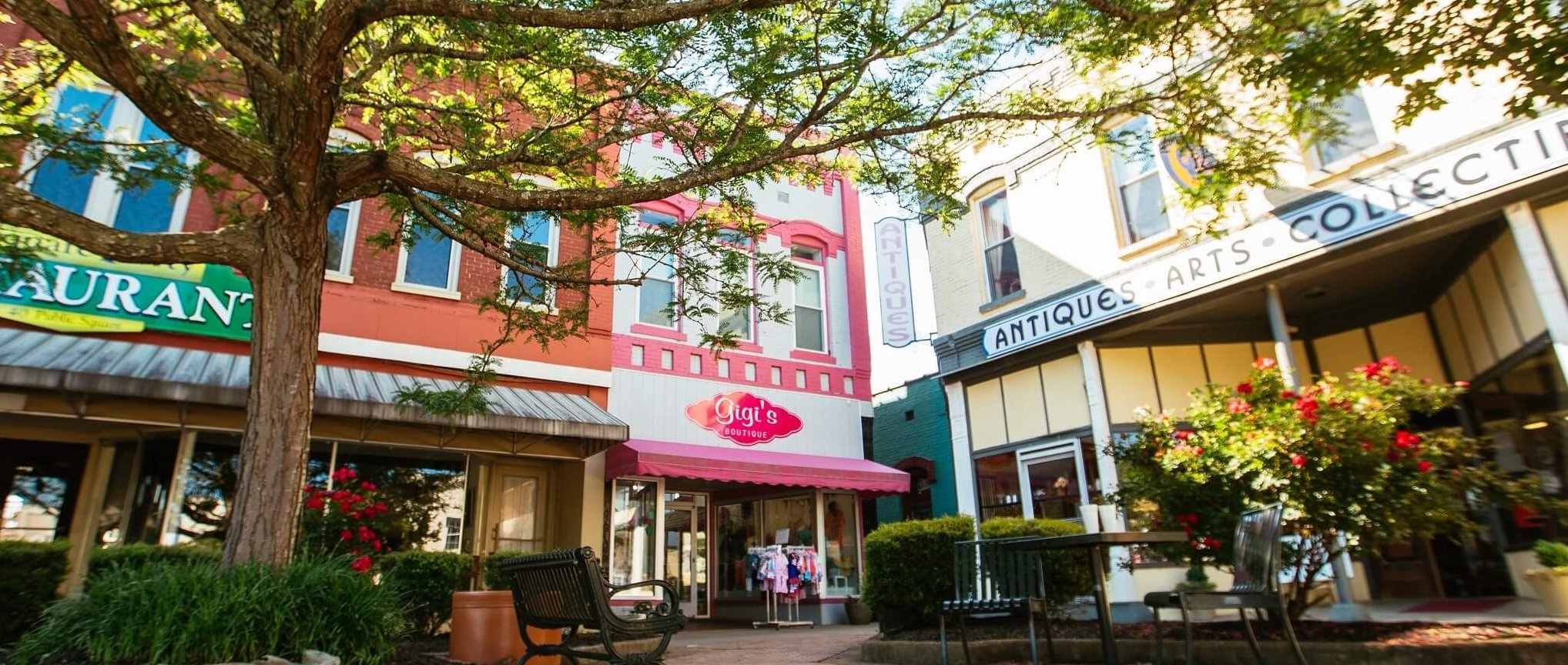 A charming downtown street with shops, including a boutique named "Gigi’s" and an antiques store, shaded by a large tree with green leaves and outdoor benches under sunny skies.