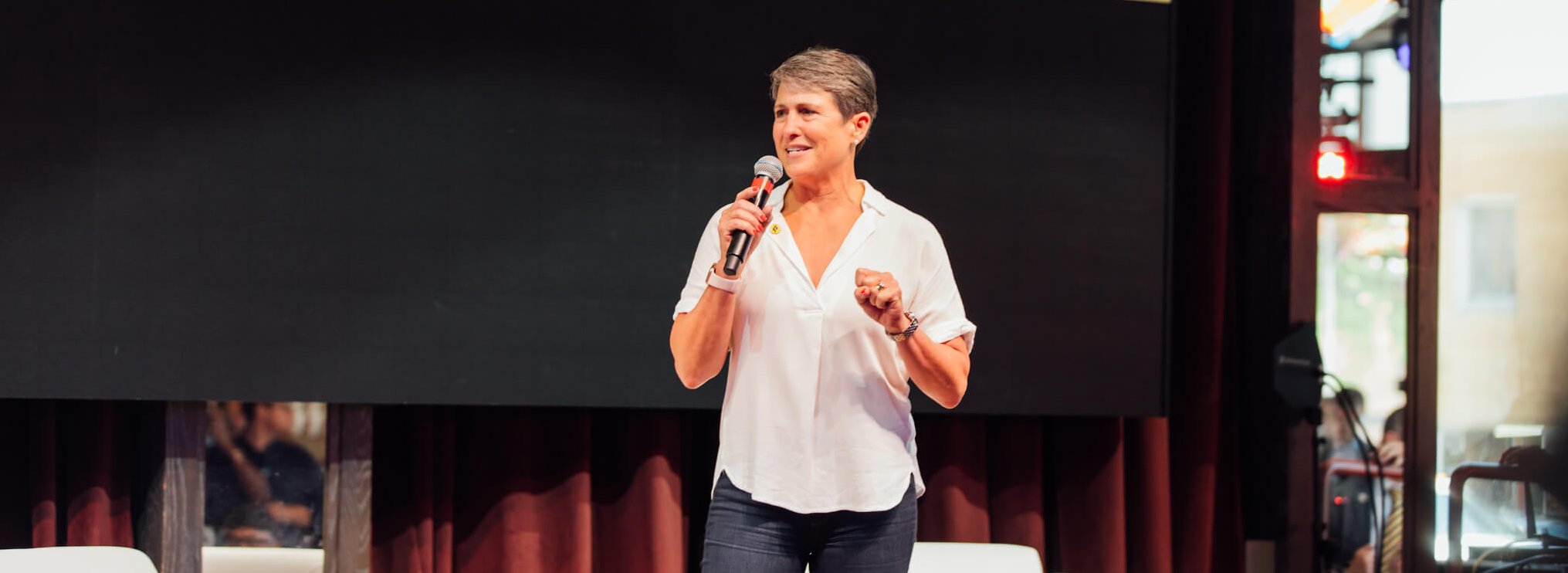 A person in a white shirt and jeans stands on a stage, holding a microphone and speaking. A large blank screen is behind them, with dark curtains and some light coming from the side.