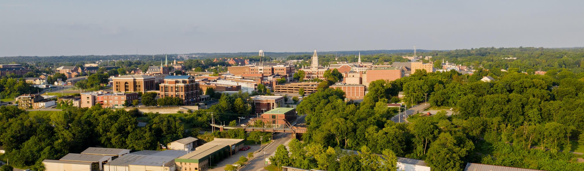 Aerial view of a small city with clusters of brick buildings, a church with a tall steeple, tree-lined streets, and surrounding green wooded areas under a clear sky.