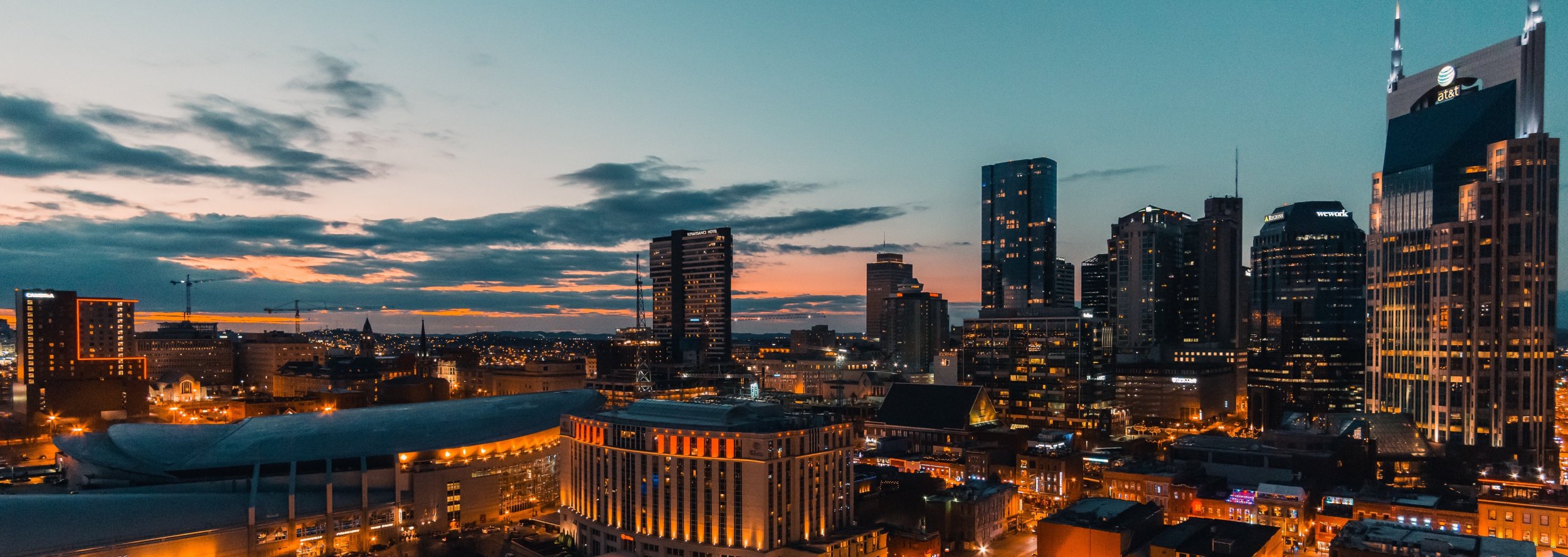 A city skyline at dusk with tall buildings illuminated by lights, a dramatic sunset with orange and blue hues in the sky, and a mix of modern and older architecture throughout the cityscape.