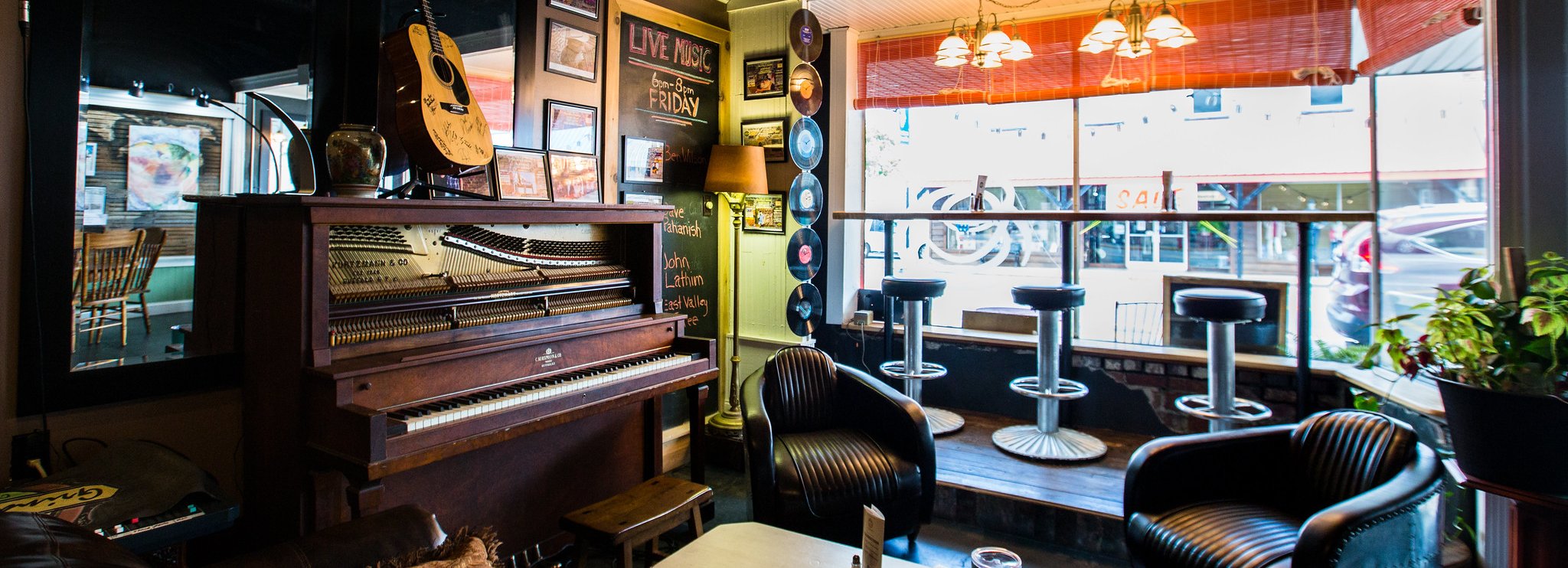Cozy cafe interior with a piano, guitar, framed photos, vinyl records on the wall, leather chairs, and barstools by a large window overlooking the street. Warm lighting creates an inviting atmosphere.