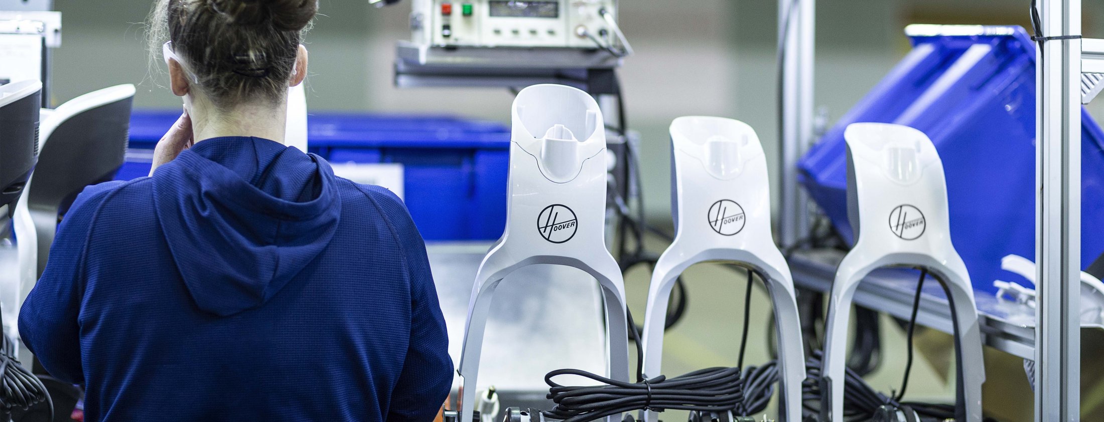 A person in a blue shirt works at an assembly line with several upright vacuum cleaner handles, each labeled with the Hoover logo, and some equipment and bins in the background.