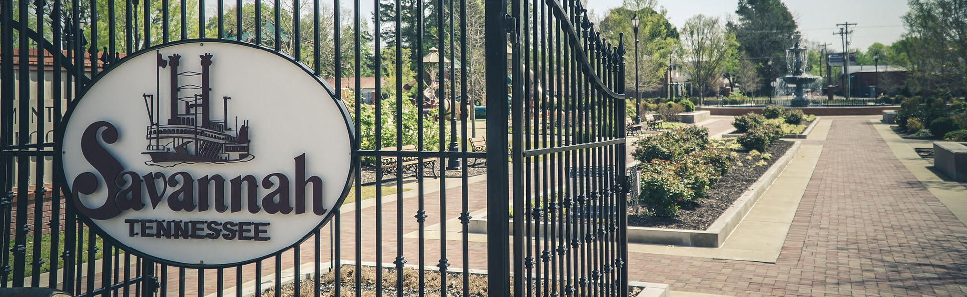 Ornamental metal gate featuring a circular sign reading "Savannah Tennessee" with a riverboat graphic, opens to a landscaped park with brick pathways, garden beds, and trees in the background.