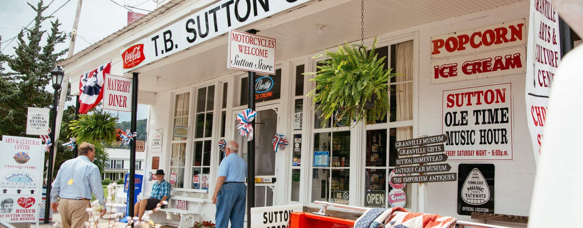 A historic country store with white wooden exterior, red and black signs for popcorn, ice cream, and music; people stand and sit outside while American flags and ferns decorate the porch.