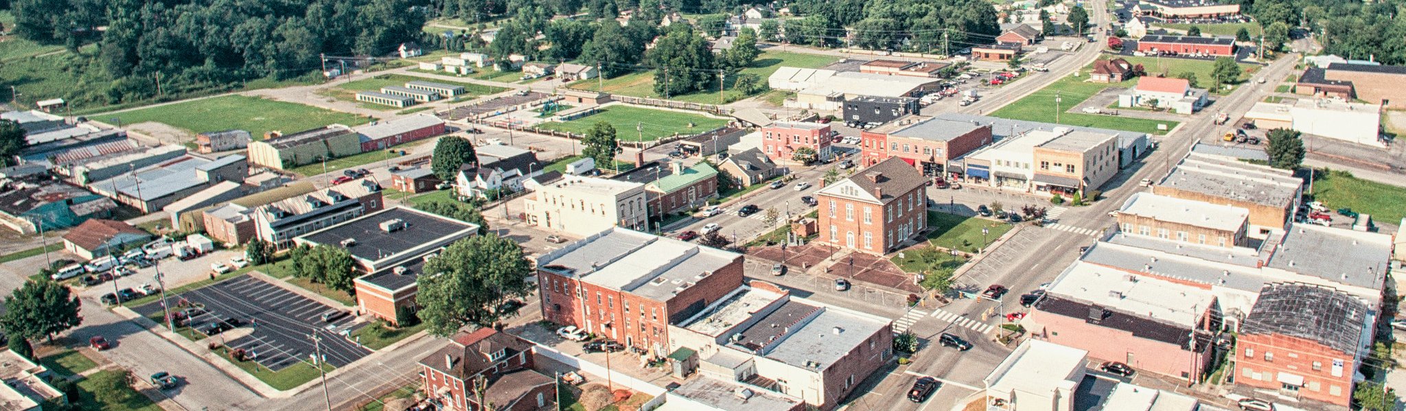 Aerial view of a small town with tree-lined streets, various brick buildings, parking lots, and patches of green spaces on a sunny day.
