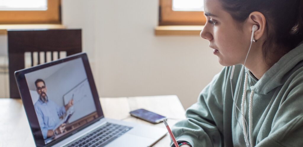A young woman wearing earphones sits at a table, watching an online lesson on her laptop. A teacher is visible on the screen, pointing at a whiteboard. A notebook and phone lie on the table nearby.