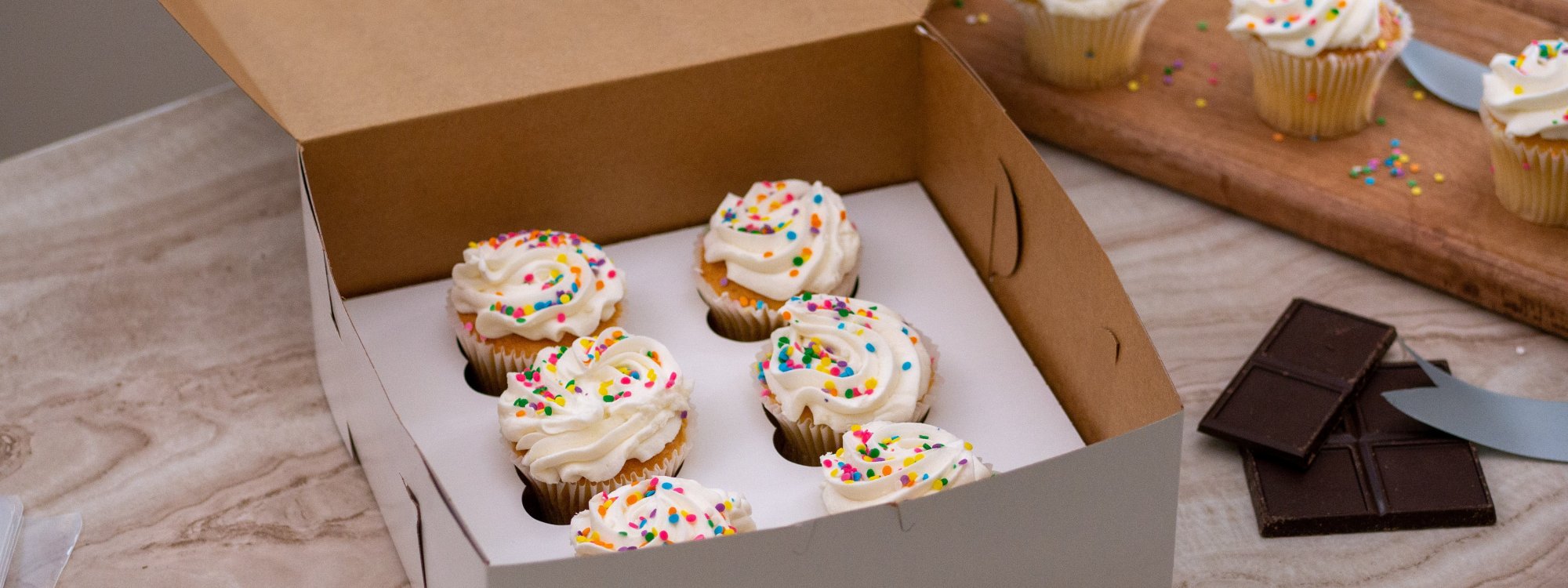 An open box with six cupcakes topped with white frosting and colorful sprinkles sits on a marble counter; additional cupcakes and chocolate pieces are nearby.