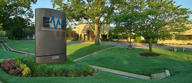 A large sign reading "EMS Engineered Medical Systems" stands on a well-manicured lawn with flowers. Behind it is a modern building surrounded by trees and landscaping, with the address 3325 on the sign.