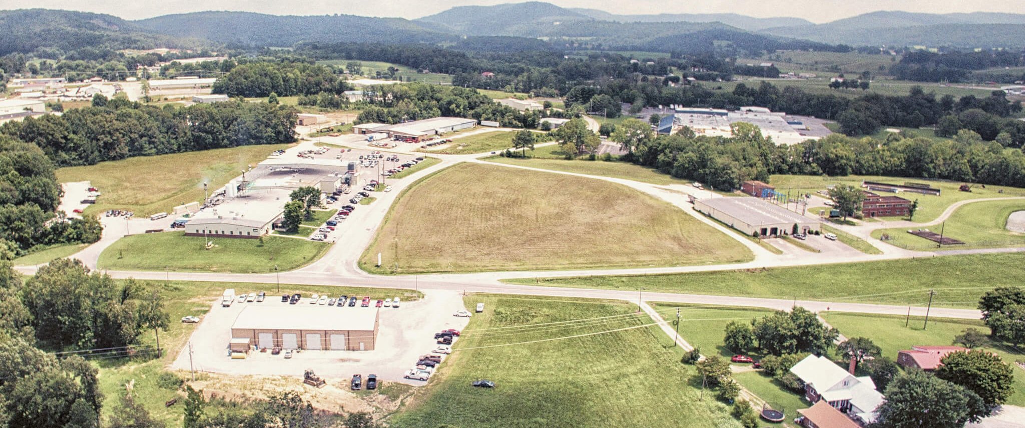 Aerial view of a small rural town with green fields, roads, scattered buildings, parking lots with cars, and tree-covered hills in the background under a partly cloudy sky.