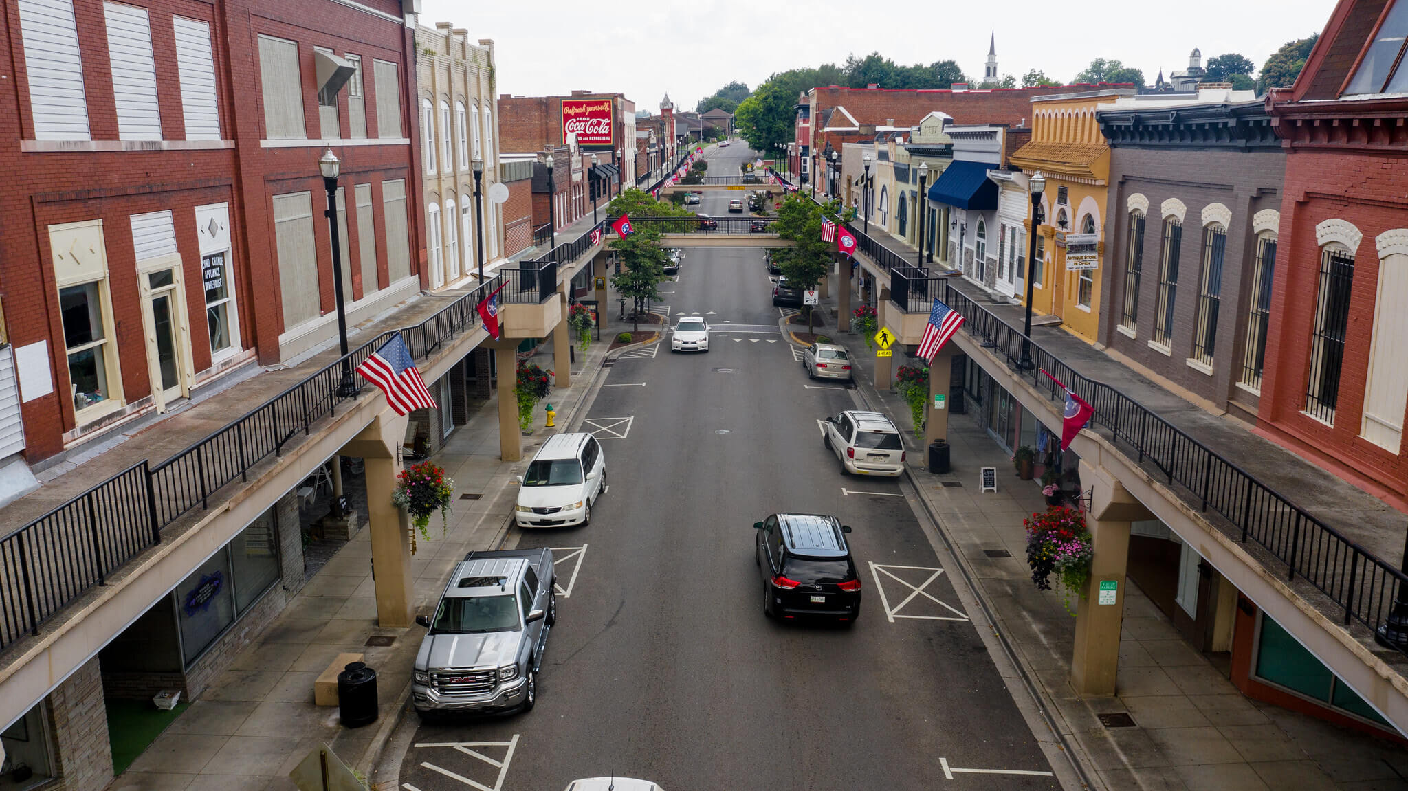 A view down a small town main street with parked cars, American flags, brick buildings, upper-level walkways, and hanging flower baskets lining both sides of the road.