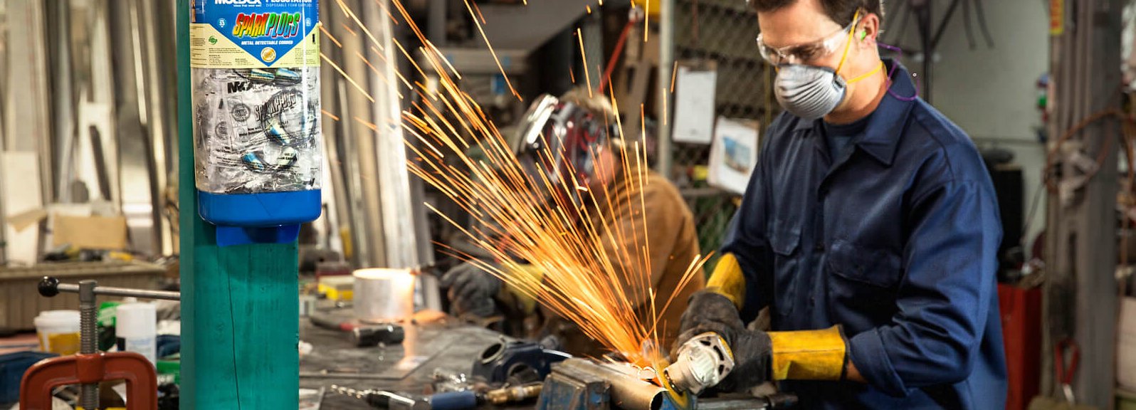 A person wearing safety gear uses an angle grinder on metal, creating a shower of sparks in a busy workshop. Another person works in the background near tools and equipment.