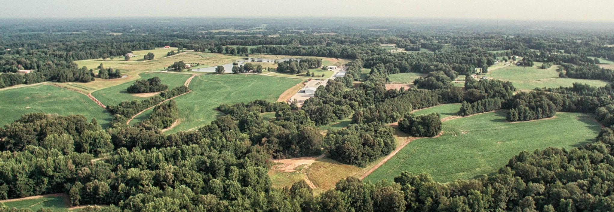 Aerial view of a large rural landscape with green fields, clusters of trees, winding dirt roads, and a few buildings scattered throughout the area under a hazy sky.