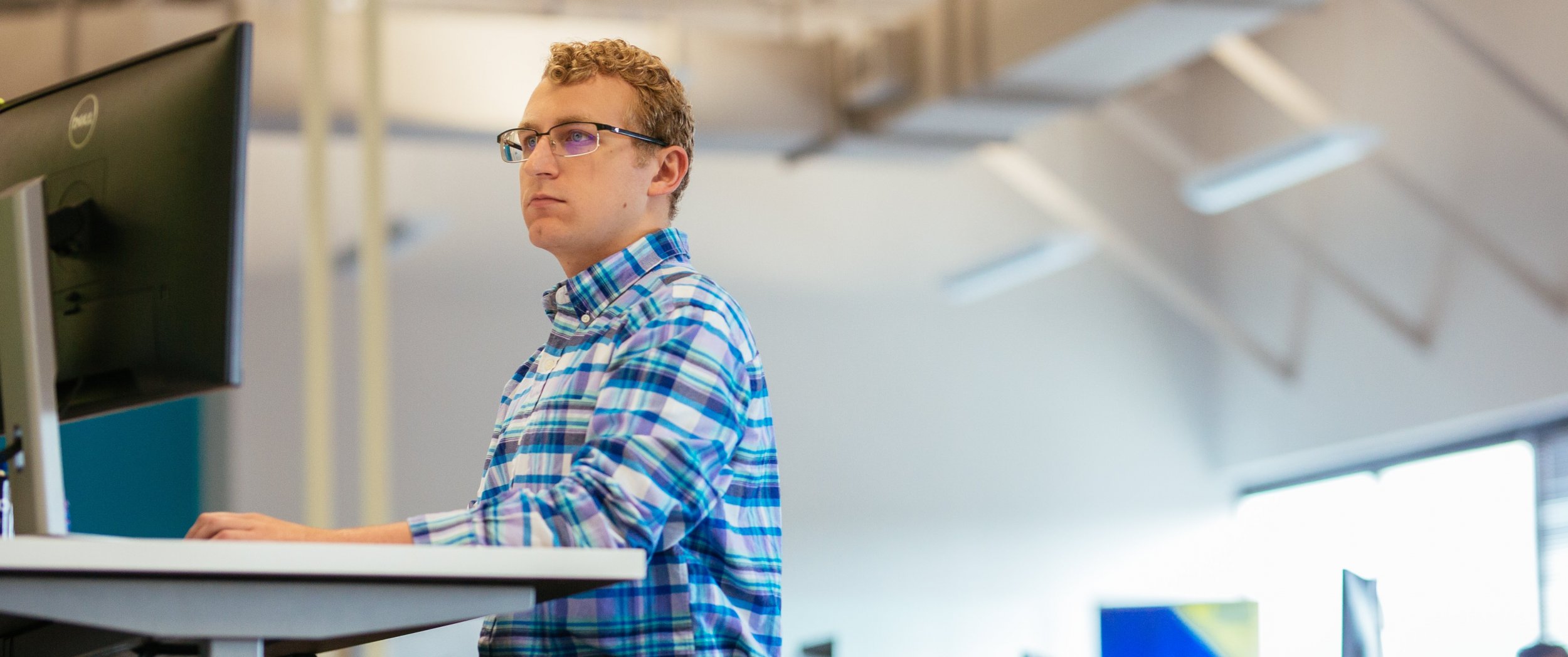 A person wearing glasses and a blue plaid shirt stands at a desk, working on a computer in a bright, modern office with high ceilings.