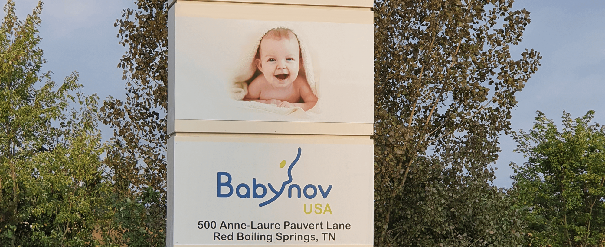 A sign for Babynov USA displays a smiling baby wrapped in a blanket above the company name and address: 500 Anne-Laure Pauvert Lane, Red Boiling Springs, TN. Trees and greenery are in the background.