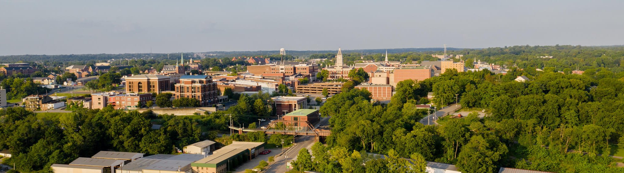 Aerial view of a small city with brick buildings, tree-lined streets, and green spaces under a clear sky on a sunny day.